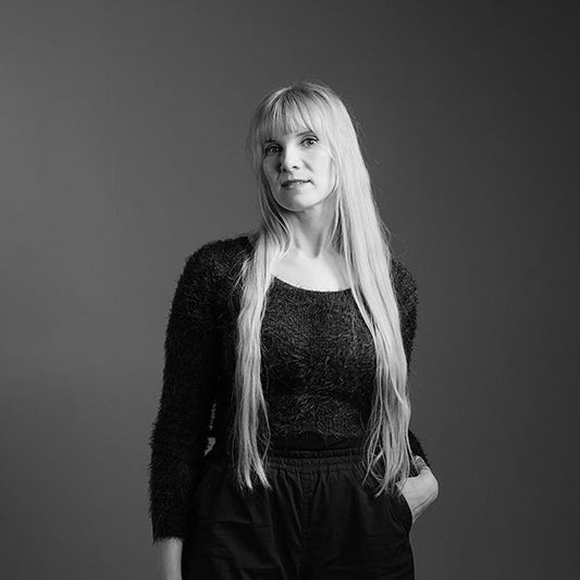 Mustavalkoinen muotokuva Katri Naukarista. Hän seisoo neutraalin taustan edessä, katse hieman ylöspäin suunnattuna. Hänellä on yllään pörröinen pitkähihainen paita ja väljät housut. Pitkät, suorat hiukset laskeutuvat hänen olkapäilleen ja selkäänsä.