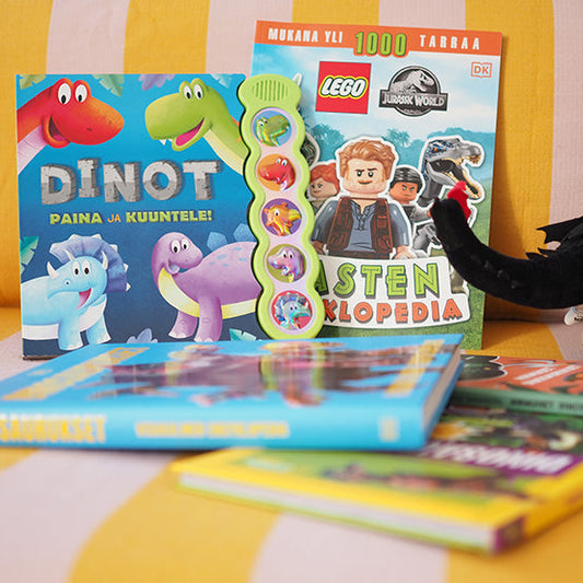 Pehmeällä sohvalla lepäävät lasten dinosauruskirjat, kuten ääninappikirja Dinot – Paina ja kuuntele!, LEGO Jurassic World -tarrakirja ja muita värikkäitä dinokirjoja. Oikeassa reunassa näkyy musta dinosauruspehmo.
