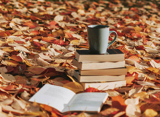 Syksyiselle maahan pudonneiden ruskanväristen lehtien peittämälle nurmikolle on asetettu kirjoja ja niiden päällä on kahvikuppi. Yksi kirja on avattuna edessä.