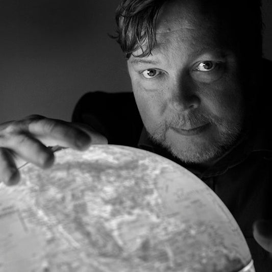 Kirjailija Markus Leikola katsoo kameraan hämärässä valaistuksessa. Hän pitää käsillään kirkkaasti valaistua karttapalloa, joka heijastaa valoa hänen kasvoilleen. Kuva on mustavalkoinen ja tunnelmallinen.