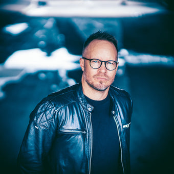 Matti Laine katsoo suoraan kameraan vakavana, yllään musta nahkatakki ja pyöreät silmälasit. Taustalla sinisävyinen, hämärä ympäristö, jossa näkyy valoa ja epätarkkoja muotoja.