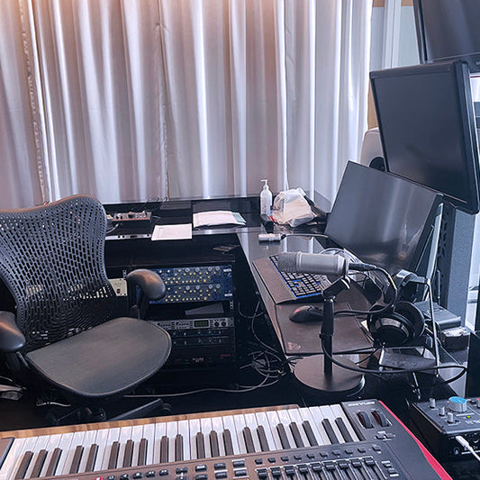 Kuvassa on äänitysstudio, jossa on työpöytä, näyttöjä, mikrofoni, kuulokkeet, koskettimisto ja työtuoli. Taustalla on verhot ja pöydän alla äänityslaitteita.