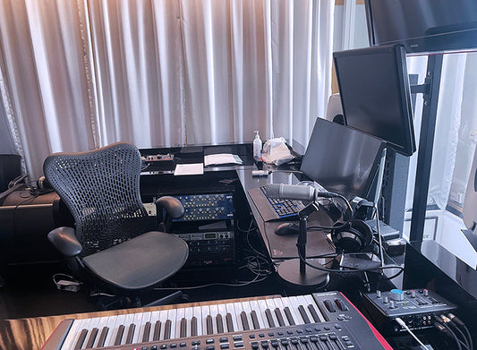 Kuvassa on äänitysstudio, jossa on työpöytä, näyttöjä, mikrofoni, kuulokkeet, koskettimisto ja työtuoli. Taustalla on verhot ja pöydän alla äänityslaitteita.