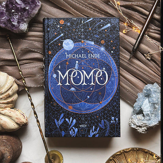 Michael Enden kirja "Momo" aseteltuna pöydälle, ympärillä esineitä kuten simpukoita, kiteitä, ametisti, kirjoitusvälineitä ja valonauha. Kirjan kannessa on tummansininen pohja, tähtikuvioita, kasviaiheita ja geometrisia elementtejä.