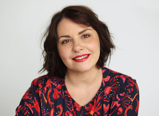 Kirjailija Clarissa Sabard katsoo kameraan ja hymyilee. Hänellä on lyhyet, ruskeat, kevyesti laineikkaat hiukset ja punainen huulipuna. Hänellä on yllään tummansininen pusero, jossa on punainen kukkakuviointi. Tausta on vaalea ja neutraali.