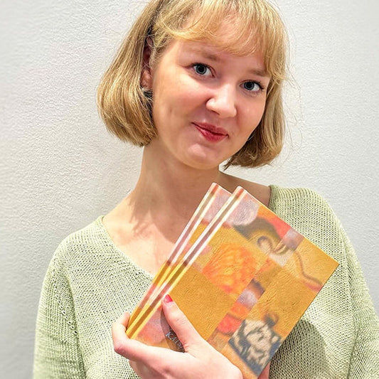 Kirjailija Kaisa Vahteristo hymyilee kameralle pitäen käsissään kirjaa Nieltyjä esineitä. Hänellä on vaaleat, polkkapituiset hiukset ja vihreä neulepaita. Taustalla näkyy vaalea seinä.