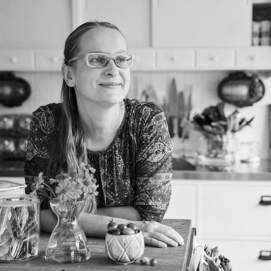 Mustavalkoisessa kuvassa ruokakirjailija Satu Koivisto istuu keittiön pöydän ääressä, nojaa käsillään pöytään ja katsoo hymyillen sivulle. Pöydällä on lasipurkkeja, yrttikasvi ja kulho marjoja. Taustalla näkyy keittiön kaappeja ja esineitä.