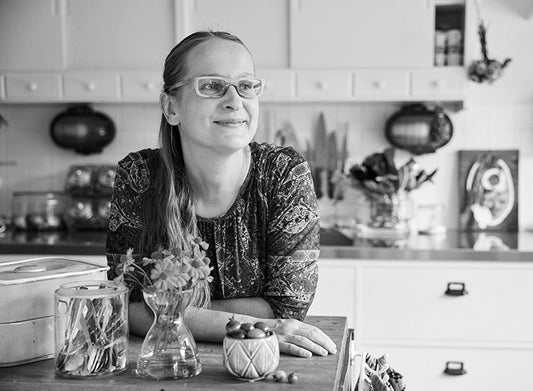 Mustavalkoisessa kuvassa ruokakirjailija Satu Koivisto istuu keittiön pöydän ääressä, nojaa käsillään pöytään ja katsoo hymyillen sivulle. Pöydällä on lasipurkkeja, yrttikasvi ja kulho marjoja. Taustalla näkyy keittiön kaappeja ja esineitä.