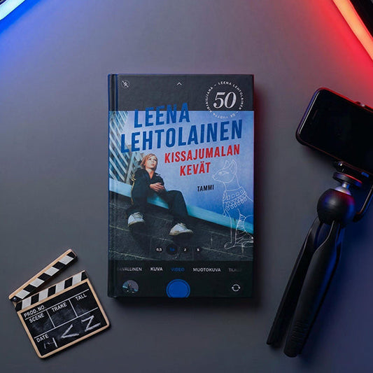 Kuvassa on Leena Lehtolaisen kirja Kissajumalan kevät. Kirjan kannen päällä näkyy puhelimen kameraa muistuttava näkymä. Kirjan vieressä on jalustassa oleva puhelin, pieni klaffitaulu sekä sinisiä ja punaisia valoputkia.