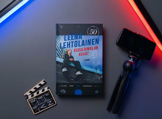 Kuvassa on Leena Lehtolaisen kirja Kissajumalan kevät. Kirjan kannen päällä näkyy puhelimen kameraa muistuttava näkymä. Kirjan vieressä on jalustassa oleva puhelin, pieni klaffitaulu sekä sinisiä ja punaisia valoputkia.