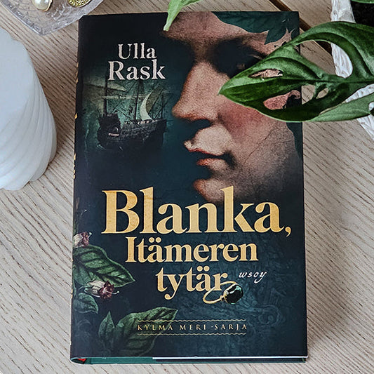 Blanka, Itämeren tytär -kirja pöydällä, ympärillä vihreä huonekasvi, valkoinen kynttilä ja sydämenmuotoinen korurasia, jossa korvakorut.