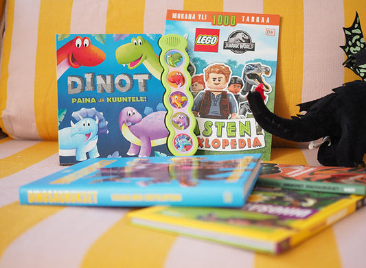 Pehmeällä sohvalla lepäävät lasten dinosauruskirjat, kuten ääninappikirja Dinot – Paina ja kuuntele!, LEGO Jurassic World -tarrakirja ja muita värikkäitä dinokirjoja. Oikeassa reunassa näkyy musta dinosauruspehmo.