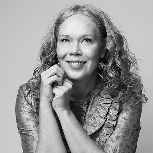 Mustavalkoinen muotokuva kirjailija Laura Lähteenmäestä. Henkilö katsoo kameraan ja hymyilee, kädet leuan alla ristissä. Hänellä on pitkät, kiharat hiukset ja kuviollinen pusero.