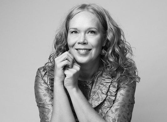 Mustavalkoinen muotokuva kirjailija Laura Lähteenmäestä. Henkilö katsoo kameraan ja hymyilee, kädet leuan alla ristissä. Hänellä on pitkät, kiharat hiukset ja kuviollinen pusero.