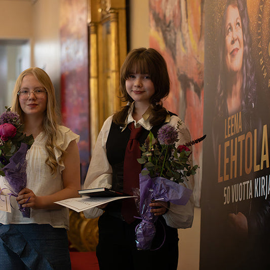Kaksi nuorta seisoo käytävällä kukkakimppujen ja todistusten kanssa. Heidän vieressään on juliste, jossa lukee “Leena Lehtolainen 50 vuotta kirjailijana”.