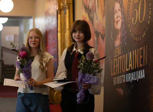 Kaksi nuorta seisoo käytävällä kukkakimppujen ja todistusten kanssa. Heidän vieressään on juliste, jossa lukee “Leena Lehtolainen 50 vuotta kirjailijana”.