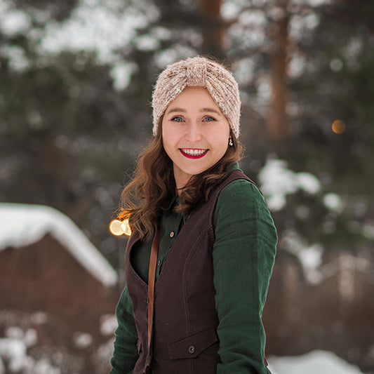 Kirjailija Lotta-Sofia Saahko hymyilee talvisessa ulkomaisemassa. Hänellä on päässään vaalea neulepanta, vihreä pusero ja ruskea liivi. Taustalla näkyy lumisia puita ja hämärtyvän illan valoja.