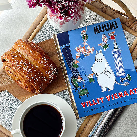 Kuvassa on tarjotin, jolla on kahvikuppi, kanelipulla puisella leikkuulaudalla, vaaleanpunaisia kukkia maljakossa sekä Tammen kultaiset kirjat -sarjan teos Muumi: Villit vieraat.