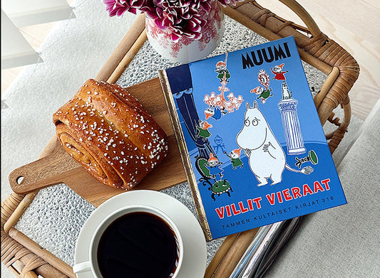 Kuvassa on tarjotin, jolla on kahvikuppi, kanelipulla puisella leikkuulaudalla, vaaleanpunaisia kukkia maljakossa sekä Tammen kultaiset kirjat -sarjan teos Muumi: Villit vieraat.