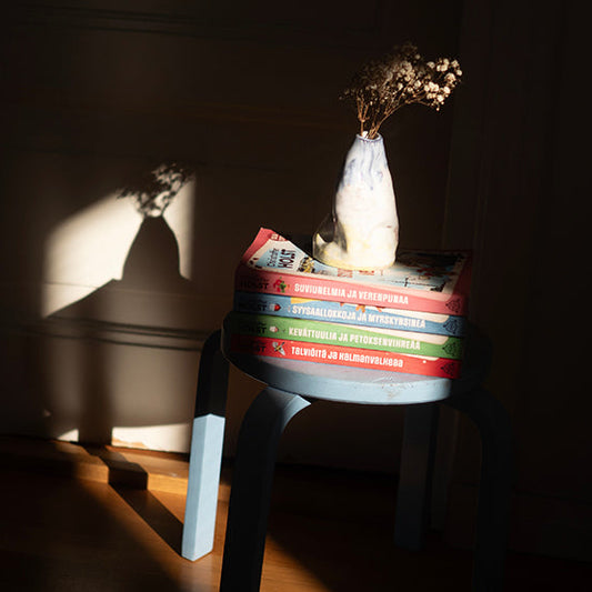 Sinisellä jakkaralla on pino värikkäitä kirjoja, joiden päällä on vaalea maljakko ja kuivakukkia. Aurinko heijastaa maljakon varjon seinälle. Kirjojen selkämyksissä näkyy dekkarien nimet eri vuodenajoista.