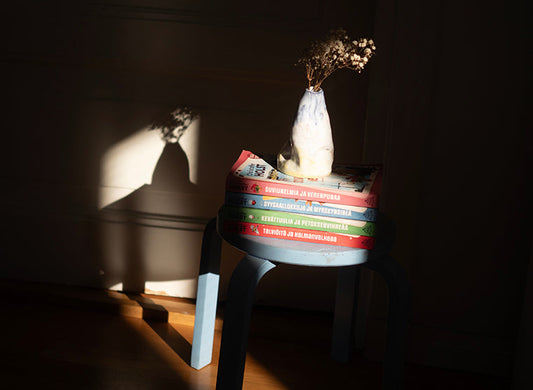 Sinisellä jakkaralla on pino värikkäitä kirjoja, joiden päällä on vaalea maljakko ja kuivakukkia. Aurinko heijastaa maljakon varjon seinälle. Kirjojen selkämyksissä näkyy dekkarien nimet eri vuodenajoista.