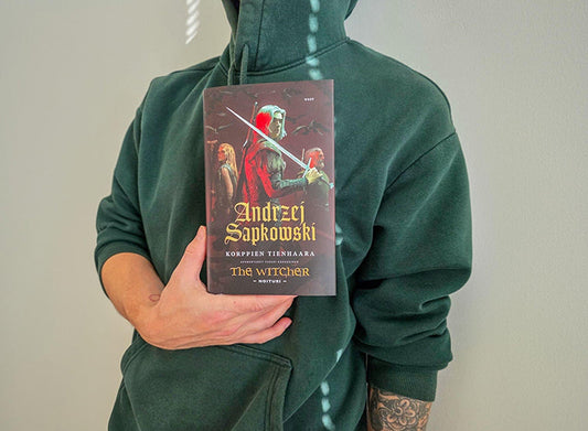 Huppariin pukeutunut henkilö pitää käsissään Andrzej Sapkowskin kirjaa ”Korppien tienhaara”. Kirjan kannessa on tumma fantasiakuva, jossa näkyy useita hahmoja ja miekka.