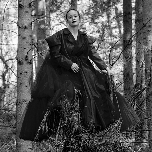 Kirjailija Olga Ravn seisoo metsässä yllään tumma, kerroksellinen mekko. Kuva on mustavalkoinen.