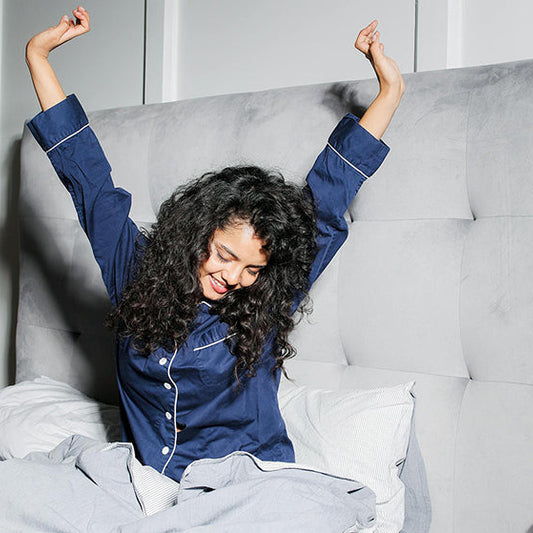 Hymyilevä nainen sinisessä pyjamassa venyttelemässä sängyllä kädet kohti kattoa.
