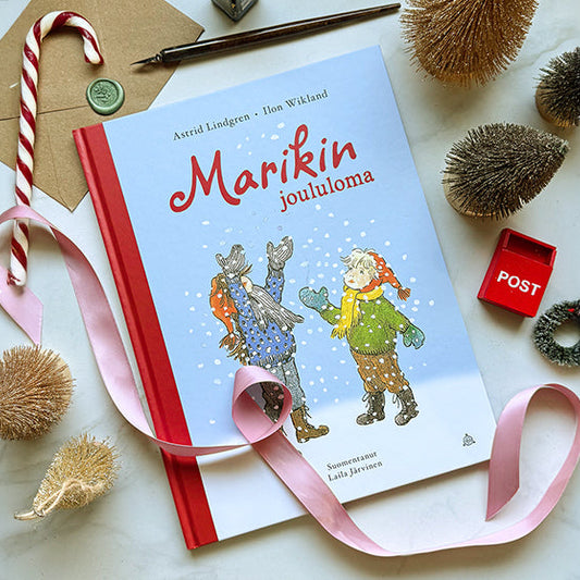 Kuvassa on Astrid Lindgrenin ja Ilon Wiklandin kirja Marikin joululoma, joka on aseteltu pöydälle vaaleanpunaisen satinauhan, karkkikepin, minikuusten, pienen punaisen postilaatikon ja kirjeaiheisten koristeiden ympäröimänä.