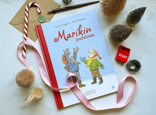 Kuvassa on Astrid Lindgrenin ja Ilon Wiklandin kirja Marikin joululoma, joka on aseteltu pöydälle vaaleanpunaisen satinauhan, karkkikepin, minikuusten, pienen punaisen postilaatikon ja kirjeaiheisten koristeiden ympäröimänä.