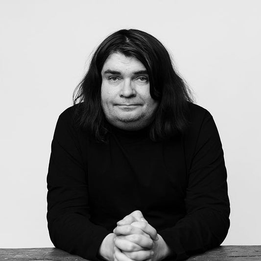 Kirjailija Samuli Knuuti istuu pöydän ääressä mustassa pitkähihaisessa paidassa. Hän katsoo kameraan, kädet lomittain edessään pöydällä. Tausta on yksivärinen ja mustavalkoinen kuva on rajattu puolikuvaksi.