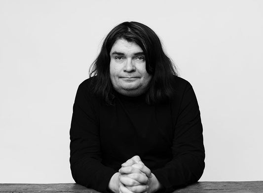 Kirjailija Samuli Knuuti istuu pöydän ääressä mustassa pitkähihaisessa paidassa. Hän katsoo kameraan, kädet lomittain edessään pöydällä. Tausta on yksivärinen ja mustavalkoinen kuva on rajattu puolikuvaksi.