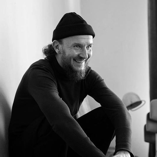 Samuli Putro istuu kyykyssä ja hymyilee leveästi katsoen sivulle. Hänellä on yllään tumma pipo ja villapaita. Kuva on mustavalkoinen, ja taustalla näkyy tuoleja ja hattu.
