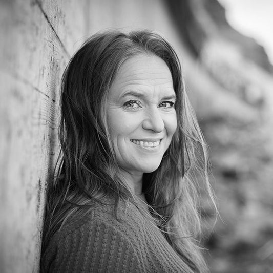 Mustavalkoinen kasvokuva kirjailija Satu Rämöstä nojaamassa kiviseinään hymyillen.