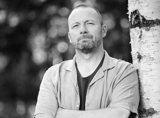 Mustavalkoinen muotokuva Teppo Heinolasta, joka nojaa koivunrunkoon kädet ristissä rinnalla. Hän katsoo suoraan kameraan vakava, rauhallinen ilme kasvoillaan. Taustalla on pehmeästi sumentunut metsämaisema.