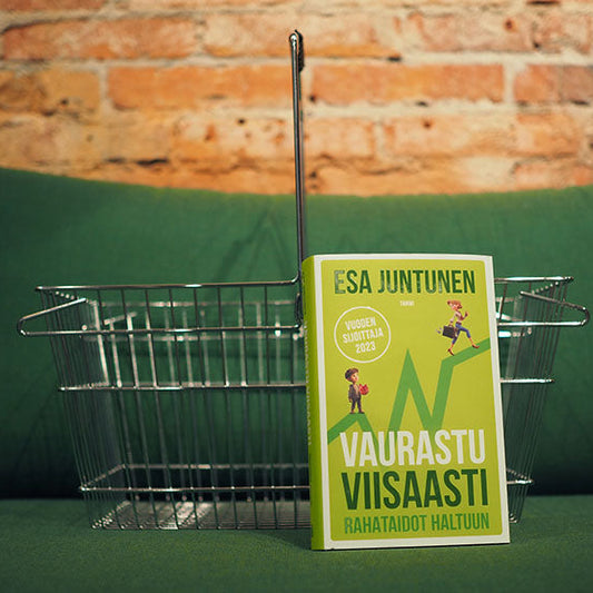 Vihreäkantinen kirja 'Vaurastu viisaasti – Rahataidot haltuun' (Esa Juntunen) nojaa metalliseen ostoskoriin, joka on asetettu vihreälle sohvalle. Taustalla näkyy tiiliseinä.