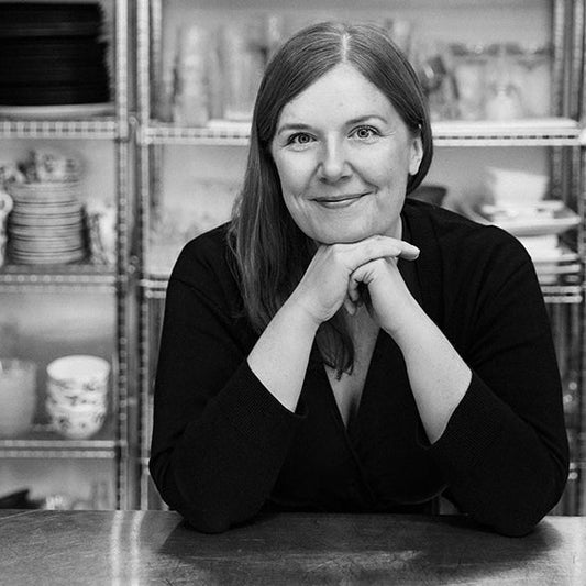 Mustavalkoinen lähikuva kirjailija Elina Innasesta, joka istuu pöydän ääressä nojaten käsiinsä hymyillen. Taustalla hyllyjä, joissa on astioita ja keittiötarvikkeita.