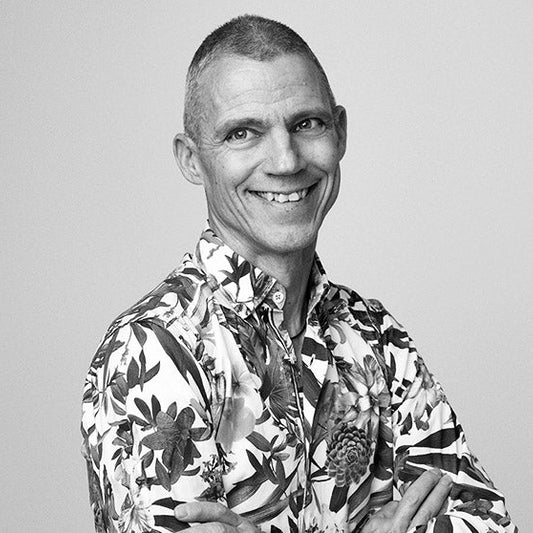 Kirjailija Jari Järvelä seisoo mustavalkoisessa lähikuvassa hymyillen ja kameraan katsoen. Hänellä on lyhyet hiukset ja yllään näyttävä kukkakuvioinen paita. 