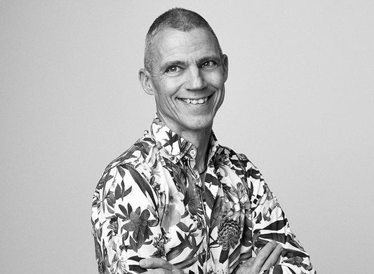 Kirjailija Jari Järvelä seisoo mustavalkoisessa lähikuvassa hymyillen ja kameraan katsoen. Hänellä on lyhyet hiukset ja yllään näyttävä kukkakuvioinen paita. 