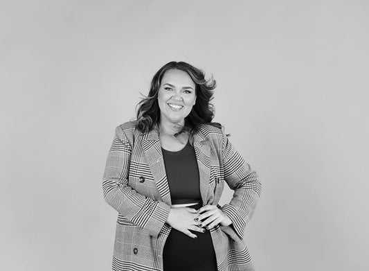 Mustavalkoinen valokuva kirjailija Veera Papinojasta. Hän hymyilee vaaleaa taustaa vasten ja hänellä on päällään ruudullinen bleiseri, kädet vatsan päällä.