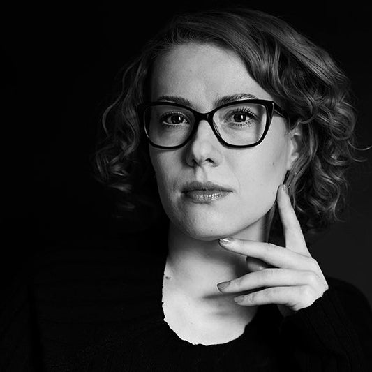 Mustavalkoinen muotokuva Laura Solasta. Hänellä on lyhyet, kiharat hiukset ja silmälasit, ja hän pitää kättään kevyesti leuallaan. Tausta on tumma.