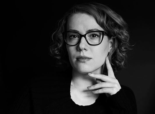 Mustavalkoinen muotokuva Laura Solasta. Hänellä on lyhyet, kiharat hiukset ja silmälasit, ja hän pitää kättään kevyesti leuallaan. Tausta on tumma.