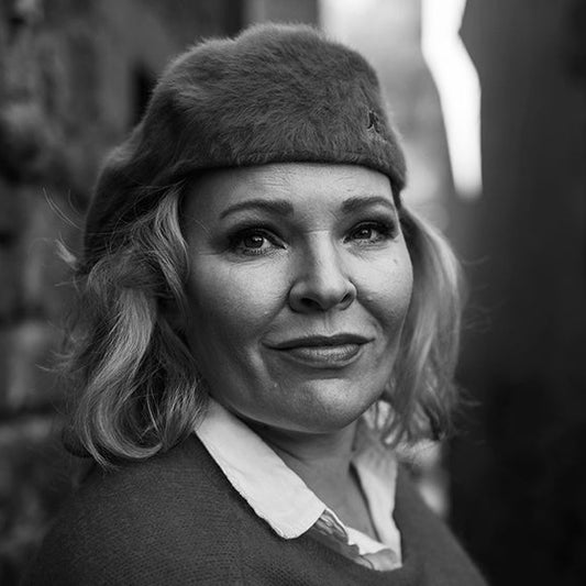 Mustavalkoinen muotokuva Milja Kaunistosta, joka katsoo suoraan kameraan. Hänellä on vaalea, hartioille ulottuva tukka ja päässään karvabaskeri. Hän pukeutuu kauluspaitaan ja neuleeseen. Taustalla kiviseinä ja kapea kuja.