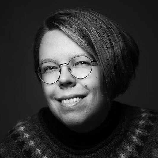 Mustavalkoisessa muotokuvassa kirjailija Paula Sankelo katsoo kameraan ja hymyilee. Hänellä on lyhyet hiukset, pyöreät silmälasit ja kuvioitu neulepusero. Tausta on tumma.