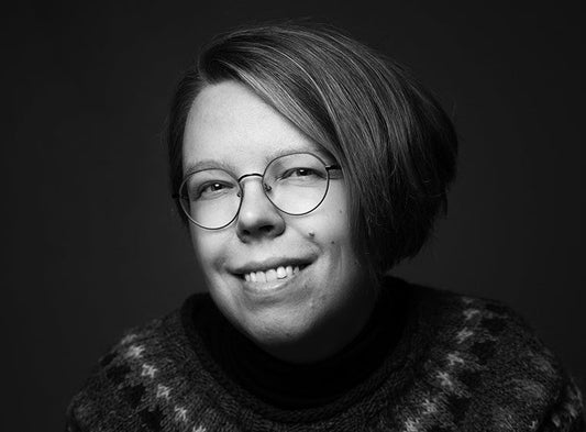 Mustavalkoisessa muotokuvassa kirjailija Paula Sankelo katsoo kameraan ja hymyilee. Hänellä on lyhyet hiukset, pyöreät silmälasit ja kuvioitu neulepusero. Tausta on tumma.