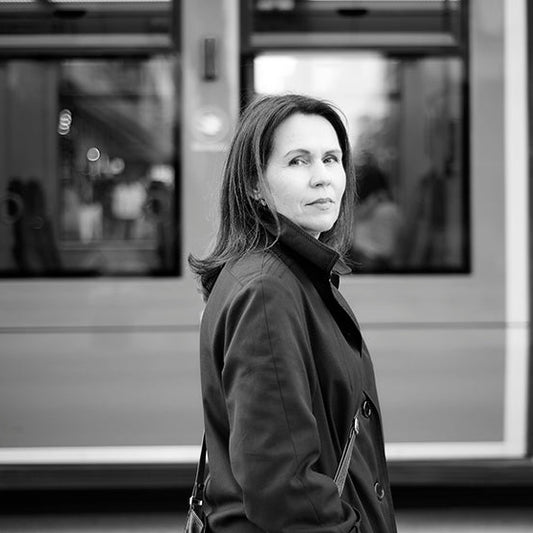 Mustavalkoisessa kuvassa kirjailija Tiina Laitila Kälvemark seisoo tummassa takissa ja katsoo kameraan. Hänen takanaan on pysäkillä oleva raitiovaunu.