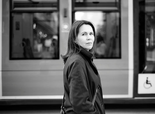 Mustavalkoisessa kuvassa kirjailija Tiina Laitila Kälvemark seisoo tummassa takissa ja katsoo kameraan. Hänen takanaan on pysäkillä oleva raitiovaunu.