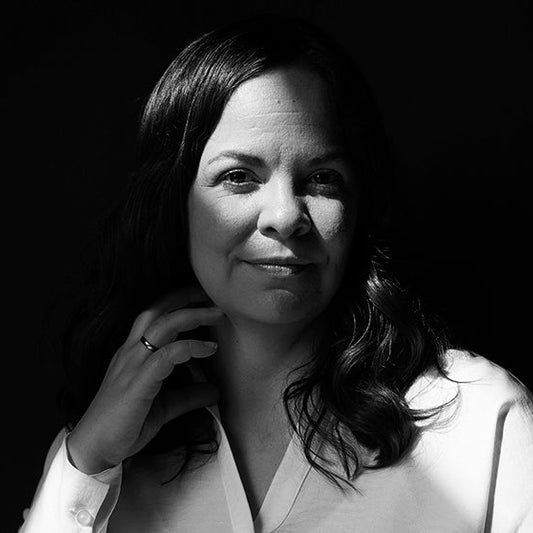 Mustavalkoinen muotokuva kirjailija Riikka Uljaksesta, joka istuu hämärässä valaistuksessa. Hänellä on keskipitkät tummat hiukset ja yllään vaalea paita. Riikka Uljas katsoo rauhallisesti kameraan, toinen käsi kevyesti kaulalla.
