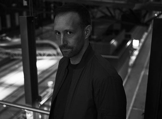 Mustavalkoisessa kuvassa kirjailija Tuomas Niskakangas katsoo kameraan hämärässä kaupunkiympäristössä. Taustalla näkyy valaistuja raiteita ja rakenteita.