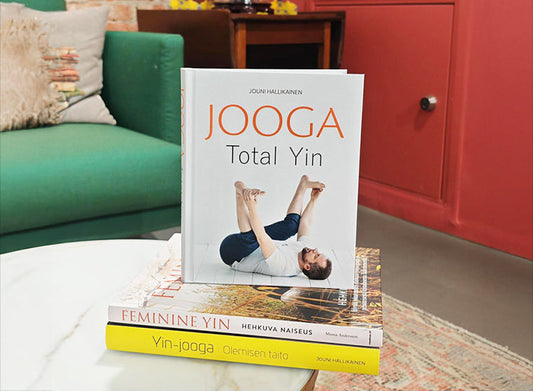 Pöydällä pinossa kolme joogakirjaa, päällimmäisenä kirja “Jooga Total Yin”, jonka kannessa on jooga-asentoa tekevä henkilö. Taustalla näkyy vihreä sohva ja punainen kaappi.
